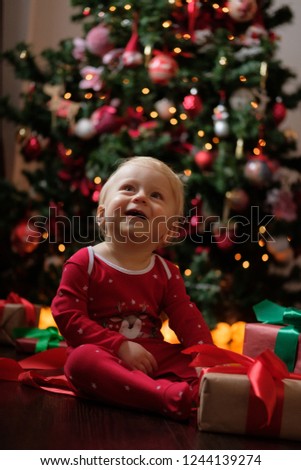 Happy baby in red pajamas on Christmas morning unpacks presents in front of the Christmas tree on a dark wooden floor