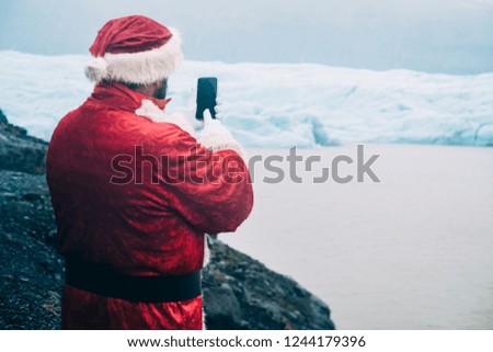 Traditional authentic style Santa Claus taking a picture his mobile phone near a glacier in Iceland