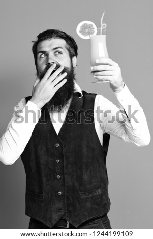 handsome bearded man with long beard and mustache has stylish hair on surprised face holding glass of alcoholic cocktail in vintage suede leather waistcoat on red studio background.