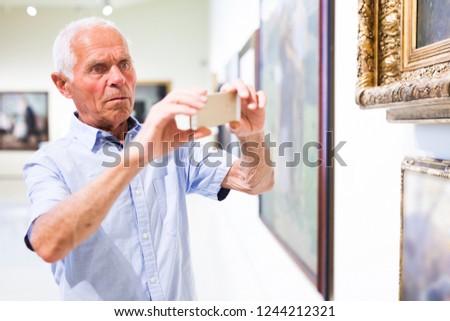 Attentive senior man taking photo by phone at painting exhibition in museum