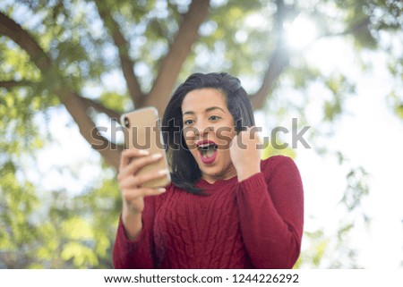 Closeup photo of casually-dressed  girl standing outdoors looking attentively and happy at screen of cellphone, winning expression raises fist up mouth opened. Received good news.