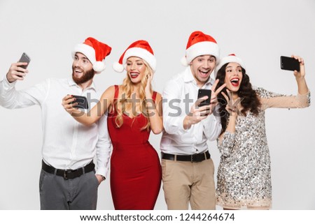 Group of happy friends dressed in red hats standing isolated over white background, celebrating New Year, taking a selfie with mobile phones