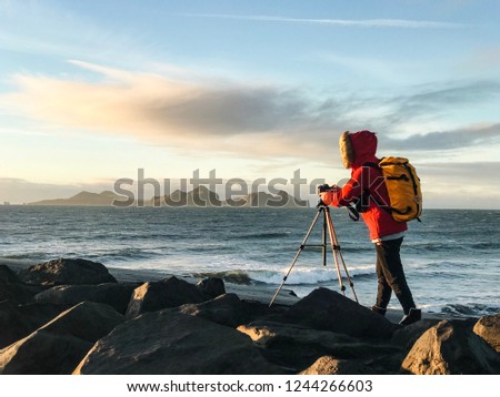 Girl in a red jacket with a yellow backpack making photos of islands on black sand beach in Iceland. Magical landscape. 