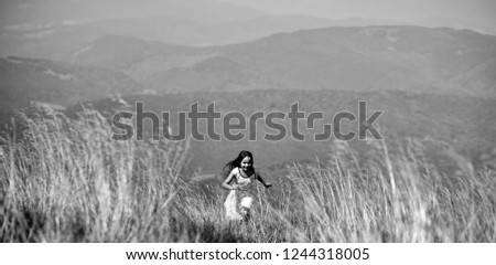 Young little pretty happy brunette girl in white lace summer dress running in mountain valley with deep dry spikelet grass in spring sunny day outdoo on natural blue background, horizontal picture
