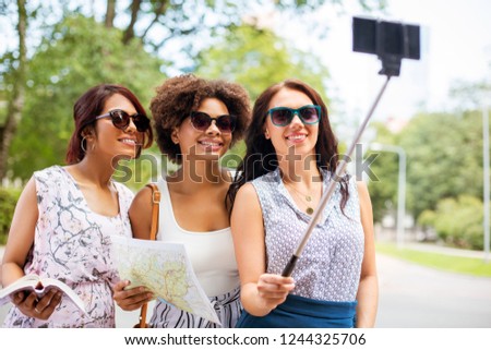 tourism, travel and technology concept - happy women with city guide and map taking picture by smartphone on selfie stick in summer park