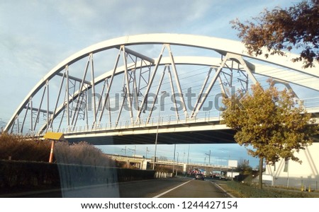 Arched steel train bridge, view from the window inside a riding car with little reflection in corner of picture from the glass.In background a road bridge. Sunny day, blue sky with some light clouds.