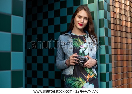 Cool funny girl model with retro film camera wearing a denim jacket, dark hair outdoors over city wall in a cage background
