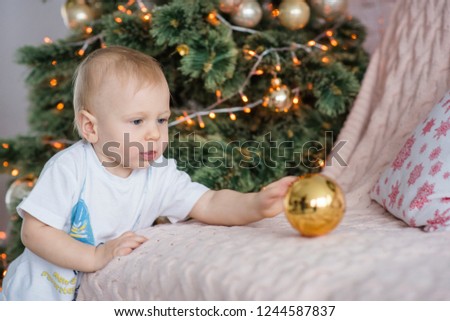Little blond boy plays with a gold ball at the Christmas tree at home. New Year holiday card. Close-up portrait of the Kid laughing, touching the toy with his finger. Waiting for a miracle. Copy space