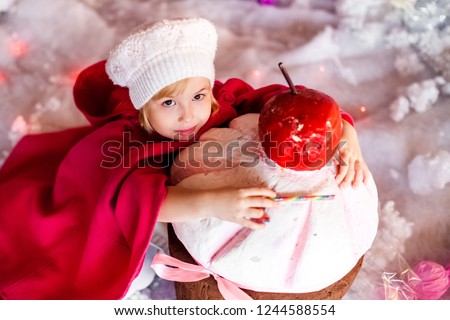 Portrait of little blonde smiling girl with white beret, red boots, pink cloak and huge artificial cake. Christmas and New Year theme.