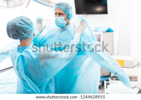 Medical workers. Portrait of nurse in sterile gloves putting on blue surgical gown on doctor