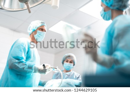 Operating room. Portrait of middle aged doctor in sterile gown holding laparoscopic instrument while assistants standing on blurred background