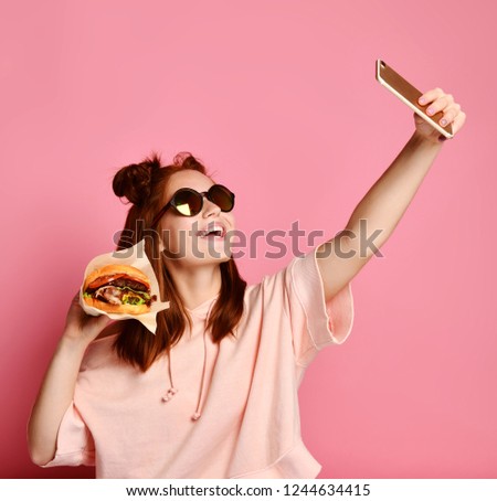 Fashion happy cool smiling girl in lifestyle clothes taking picture makes self portrait on smartphone with burger sandwich over pink background wearing sunglasses and pink jacket