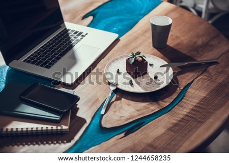 Laconic image of wooden table with lovely cake placed on the plate and modern laptop with notebooks and smartphone next to it