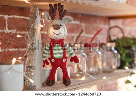 Close up toy amigurumi deer in striped sweater and stylish red butterfly tie stands on the wooden shelf near the decorated christmas tree and christmas lights.