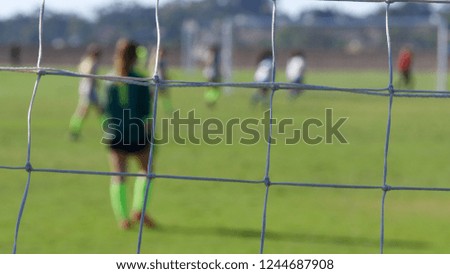 Horizontal composition of a soccer goalie through goal net in focus with shallow depth of field players in background.
