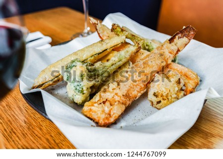 Tempura broccoli, asparagus, zucchini on grease proof paper in a black bowl on a wooden table next to glasses of red wine. Royalty-Free Stock Photo #1244767099