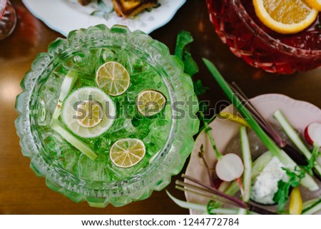 Flat lay of fruit punch in a glass punch bowl on a wooden table next to glasses of wine and cups of tea.