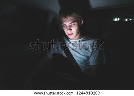 Bored and lonely teenager sitting in the back of a car on his smartphone. The light from the screen is illuminating his face.