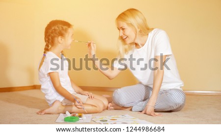 Charming girls sitting on floor with paper and watercolor and painting noses to each other having fun.