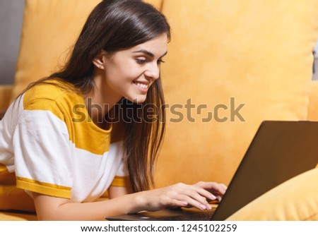 Cheerful long haired brunette girl laying with her laptop on the yellow sofa in the light living room while chatting on facebook, modern technology, communication concept