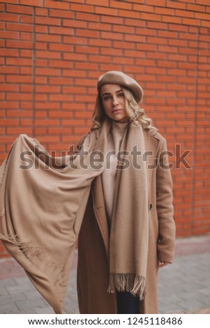 Attractive curly blonde woman dressed beret, coat, scarf and sweatshirt. Total beige or brown look. Background brick wall. Street fashion for spring, winter or fall season. Minimalistic look.