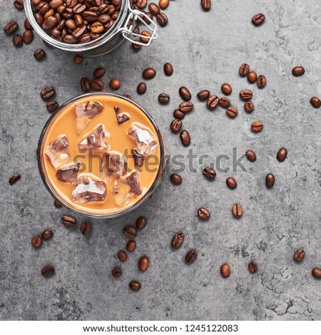 Chocolate, vanilla, caramel or cinnamon iced coffee in tall glass. Cold brewed iced coffee in glass and coffee beans in glass jar on grey concrete background. Top view, square crop.
