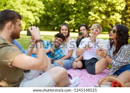 friendship, leisure and technology concept - man with camera photographing his friends drinking non alcoholic drinks at picnic in summer park