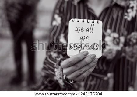 closeup of a young woman outdoors showing a notepad in front of her face with the text we all are migrants written in it, in black and white
