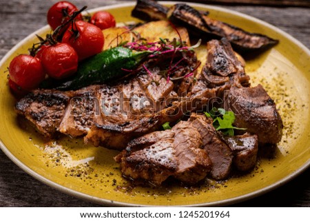 Grilled T-bone steak seasoned with spices and fresh herbs served on a wooden board with fresh tomato and roast potatoes