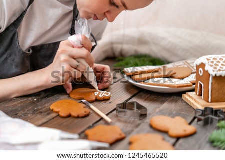 A young girl decorates ginger cookies Christmas winter morning. Woman draws Icing on honey gingerbread cookies. Wooden brown table. copy space. Blank biscuit gingerbread house, ready to decorate.