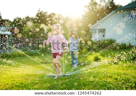 Adorable little girls playing with a sprinkler in a backyard on sunny summer day. Cute children having fun with water outdoors. Funny summer games for kids.