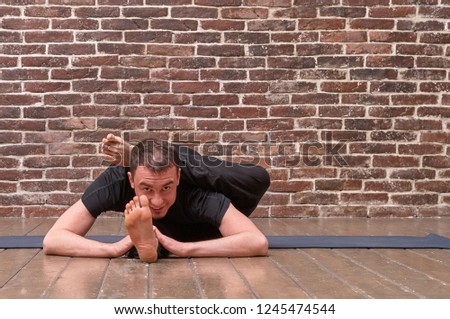 Sporty attractive young man working out, yoga, pilates, fitness training, asana Eka pada shirshasana over brick wall background. Studio shot. Full length