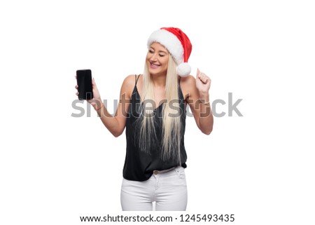 Surprised blond woman in black blouse and christmas santa hat shows a finger on a smartphone in her hand and looking at the camera over isolated white background. Sale concept