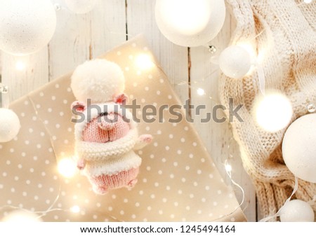 Handmade knitted toy. Christmas pig in a white sweater and hat with a large pompom on the light background of christmas balls  and lights