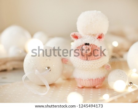 Handmade knitted toy. Christmas pig in a white sweater and hat with a large pompom on the light background of christmas balls  and lights