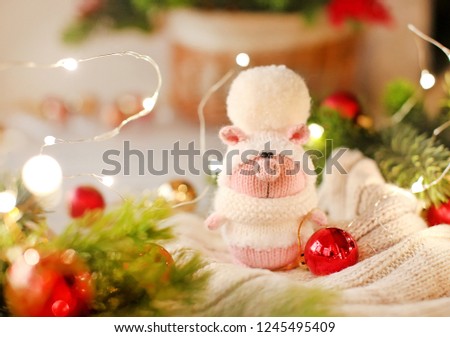 Handmade knitted toy. Christmas pig in a white sweater and hat with a large pompom on the light background of christmas red balls, branches  and lights