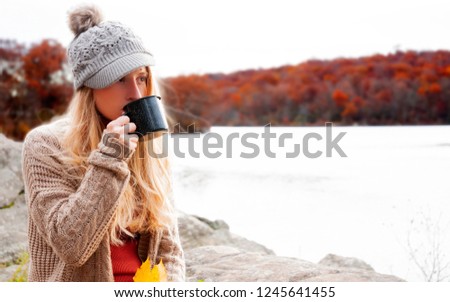 Autumn nature landscape. Woman is drinking hot tea outdoor. Beautiful woman is sitting on stone and looking at lake