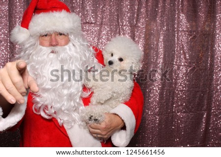 Santa Claus with a Dog. Santa holds a small white Bichon Frise dog in a Photo Booth with a Pink Sequin Backdrop. Santa loves dogs. 