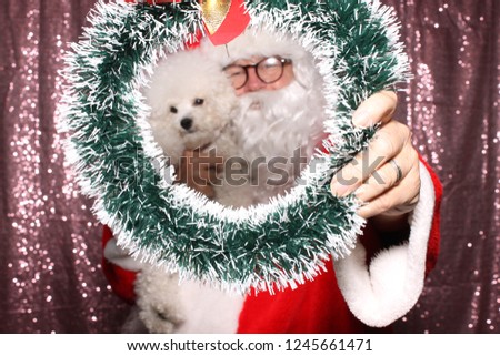 Santa Claus with a Dog. Santa holds a small white Bichon Frise dog in a Photo Booth with a Pink Sequin Backdrop. Santa loves dogs. 