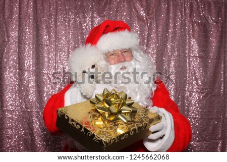 Santa Claus with a Dog. Santa holds a small white Bichon Frise dog with a gold Christmas Present in a Photo Booth with a Pink Sequin Backdrop. 