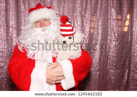 Santa Claus with a Dog. Santa holds a small white Bichon Frise dog wearing a Red and White striped hat in a Photo Booth with a Pink Sequin Backdrop. 
