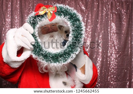 Santa Claus with a Dog. Santa holds a small dog and peers through a Christmas Wreath while in a Photo Booth with a Pink Sequin Backdrop.