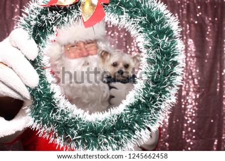 Santa Claus with a Dog. Santa holds a small dog and peers through a Christmas Wreath while in a Photo Booth with a Pink Sequin Backdrop.