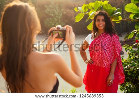 happy and beautiful indian woman in traditional stylish red sari holding the camera in hand posing for the photographer on the beach tropical paradise sea ocean