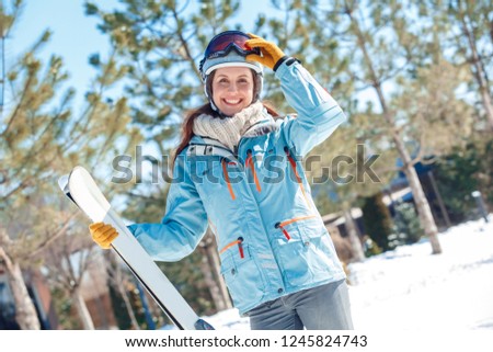 Young woman wearing helmet and goggles on a winter vacation standing outdoors holding skis looking camera smiling cheerful