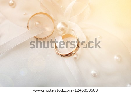 Two gold wedding engagement rings on a white pad with a bow and beads, close-up view from the top, warm colors