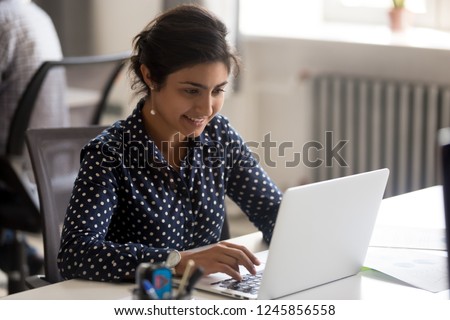 Smiling Indian female employee using laptop at workplace, looking at screen, focused businesswoman preparing economic report, working online project, cheerful intern doing computer work, typing Royalty-Free Stock Photo #1245856558