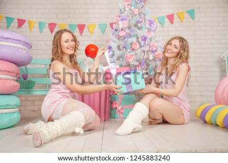 two cute best friends sit in pink pajamas on the floor, enjoy and laughing near the Christmas tree with gifts