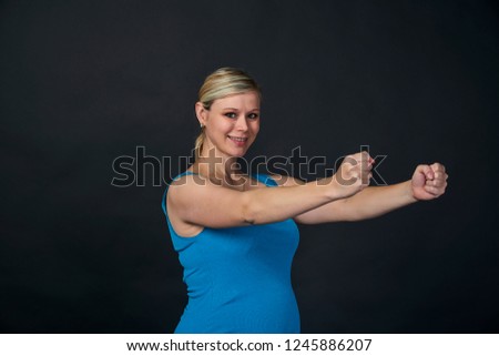 Young blondy pregnant woman in blue tank top does exercises, black background