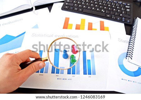 A young man is holding a magnifying glass in his hand, looking at statistic figures - a business concept on the subject of sampling and selecting the right target group and examining it Royalty-Free Stock Photo #1246083169